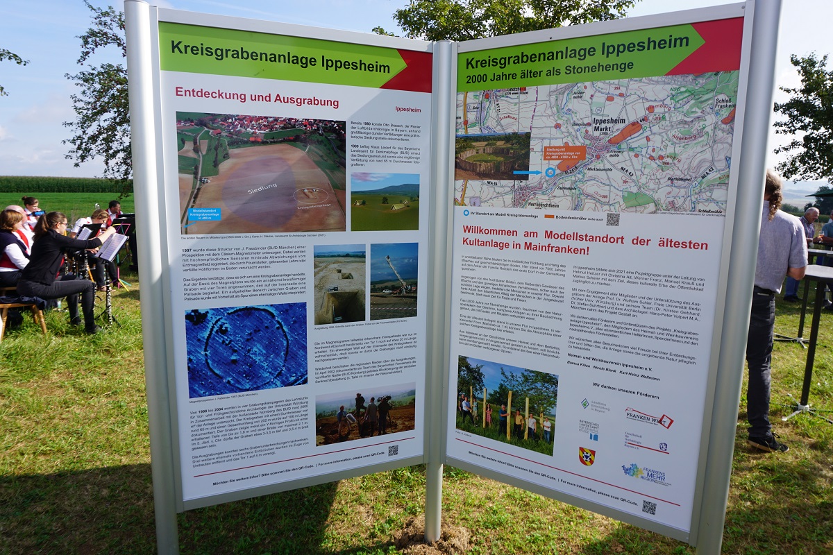 Zwei Infotafeln zur Kreisgrabenanlage Ippesheim auf Wiese bei öffentlicher Veranstaltung – linke Tafel zeigt Luftbild und Ausgrabungsfotos, rechte Tafel betont Alter der Anlage mit Lageplan und Begrüßungstext, Szene vermittelt archäologische Bedeutung und regionale Vermittlung.