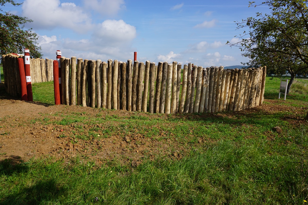 Rekonstruierte Palisadenanlage aus senkrechten Holzstämmen auf grasigem Gelände – zwei rot-weiß gestreifte Markierungspfähle am Eingang, umgeben von Feldern und Bäumen unter teils bewölktem Himmel, Szene zeigt historische Einfriedung in ländlicher Umgebung.
