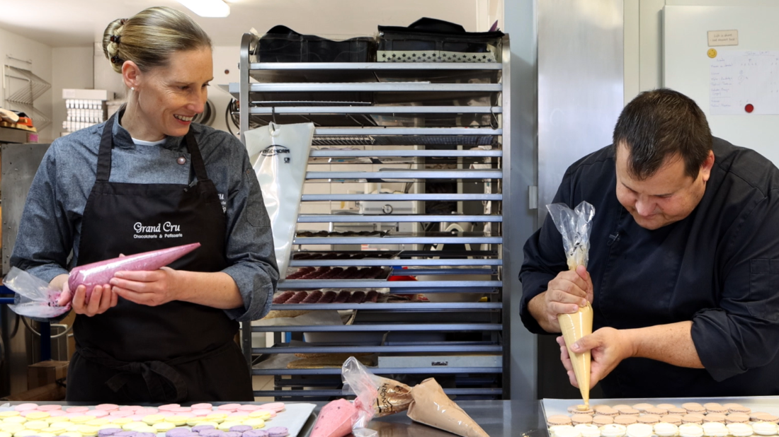 Zwei Konditoren arbeiten in einer Backstube; einer hält einen Spritzbeutel mit rosa Creme, der andere trägt gelbe Creme auf runde Formen auf – im Hintergrund ein Regal mit Backwaren und Küchengeräten.