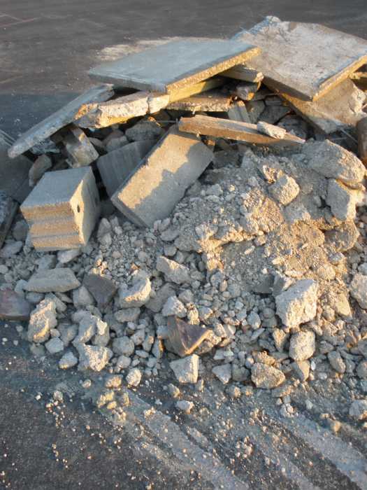 Ein Haufen aus zerbrochenem Beton mit großen Platten und kleineren Bruchstücken liegt auf dunklem Asphalt, vermutlich an einer Baustelle oder einem Abrissort.