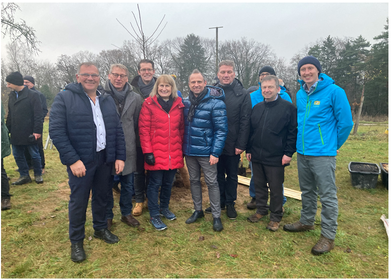 Personen in Winterkleidung stehen auf einer Wiese vor einem frisch gepflanzten jungen Baum – umgeben von kahlen Bäumen, Werkzeugen und Materialien wie Eimern und Holzlatten, deutet auf gemeinschaftliche Pflanzaktion bei kalter Witterung.