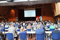 Großer Konferenzsaal mit Publikum an Tischen, Blickrichtung zur Bühne mit Podium und Präsentationsleinwand – Folientext nennt Jörg Asmussen (Deutsche Bundesbank) zur Begrüßung und Thomas Kemmerich für den Eröffnungsvortrag; Flaschen und Gläser auf den Tischen, Teilnehmer aufmerksam, Raum mit Holzverkleidung und hoher Decke.