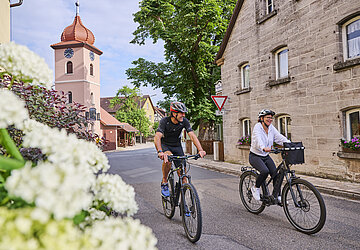 Zwei mittelalte Radfahrer im Innenort von Ditersheim mit einem Steinhaus und einem Kirchturm im Hintergrund.