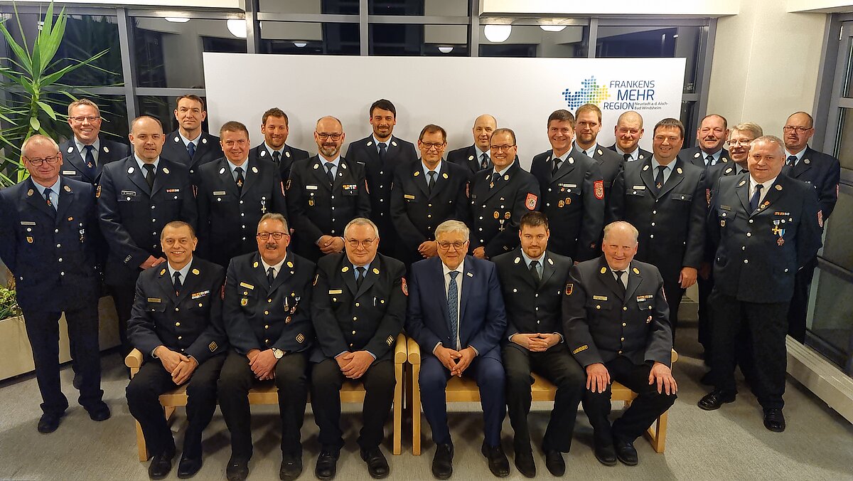 Gruppenfoto von Personen in dunklen Uniformen mit Abzeichen, die in zwei Reihen vor einem weißen Hintergrund mit dem Logo „Frankens MEHR Region“ stehen, aufgenommen in einem Innenraum mit großen Fenstern und einer grünen Pflanze links.