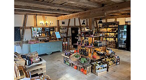 Ein liebevoll gestalteter Hofladen mit rustikalem Charme: Holzbalken, Steinboden und sorgfältig arrangierte Regale voller regionaler Produkte – von frischem Gemüse über Öle bis hin zu Feinkost. Die Kombination aus traditioneller Architektur und moderner Präsentation schafft eine warme, einladende Atmosphäre, die zum Stöbern und Genießen einlädt. Ein echtes Stück gelebter Regionalität.