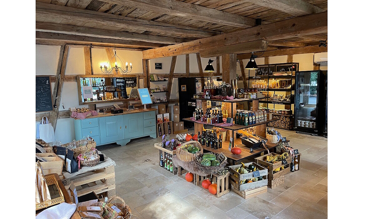 Ein liebevoll gestalteter Hofladen mit rustikalem Charme: Holzbalken, Steinboden und sorgfältig arrangierte Regale voller regionaler Produkte – von frischem Gemüse über Öle bis hin zu Feinkost. Die Kombination aus traditioneller Architektur und moderner Präsentation schafft eine warme, einladende Atmosphäre, die zum Stöbern und Genießen einlädt. Ein echtes Stück gelebter Regionalität.