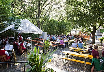 Unter Bäumen sitzende Personen auf einem Fest wo vorne dran eine Blaskapelle spielt 