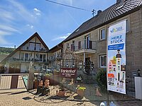 Innenhof mit dekorativem Metallbogen und Schriftzug „Ferienhof der Familie Schnell mit Udo“, flankiert von zwei Gebäuden mit Balkonen; rechts Banner mit Text „MEHR ALS MEHRWERT – HERZ STÜCK – Jeden Leben findet innen statt“ vor einladender Kulisse.