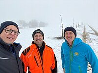 Drei Männer in Winterkleidung stehen vor einer verschneiten Landschaft mit jungen Bäumen entlang eines Weges – mittig eine orange STIHL-Jacke, rechts eine blaue Jacke mit Sonnen-Logo, im Hintergrund Nebel und Schnee, deutet auf eine Außeneinsatz- oder Pflanzaktion bei winterlichen Bedingungen.