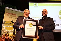 Zwei Männer stehen auf Bühne und halten gerahmte Urkunde zum „Steigerwald Panoramaweg“ – links im Anzug, rechts in schwarzem Hemd; Hintergrundbanner und Bildschirm verweisen auf die Auszeichnung „Wanderbares Deutschland – Qualitätsweg“ im Jahr 2023.