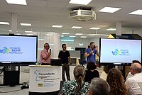 Drei Personen stehen vor zwei großen Bildschirmen mit dem Logo „Frankens MEHR Region“ und einem Tisch mit einem Banner „Gesundheitsregionplus Landkreis Neustadt a.d.Aisch-Bad Windsheim“, während eine Person ein Mikrofon hält und spricht; im Vordergrund sitzen mehrere Personen und hören zu.