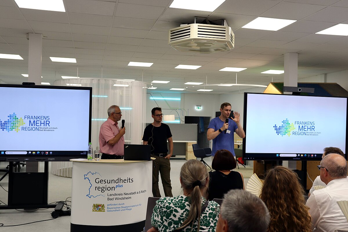 Drei Personen stehen vor zwei großen Bildschirmen mit dem Logo „Frankens MEHR Region“ und einem Tisch mit einem Banner „Gesundheitsregionplus Landkreis Neustadt a.d.Aisch-Bad Windsheim“, während eine Person ein Mikrofon hält und spricht; im Vordergrund sitzen mehrere Personen und hören zu.