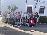 Eine fröhliche Gruppe versammelt sich auf dem Kopfsteinpflaster vor einem hellblauen Haus mit Weinmotiv – Gläser erhoben, Kronen auf dem Kopf und ein Hauch von Festlichkeit in der Luft. Ein Moment voller Gemeinschaft und Genuss.