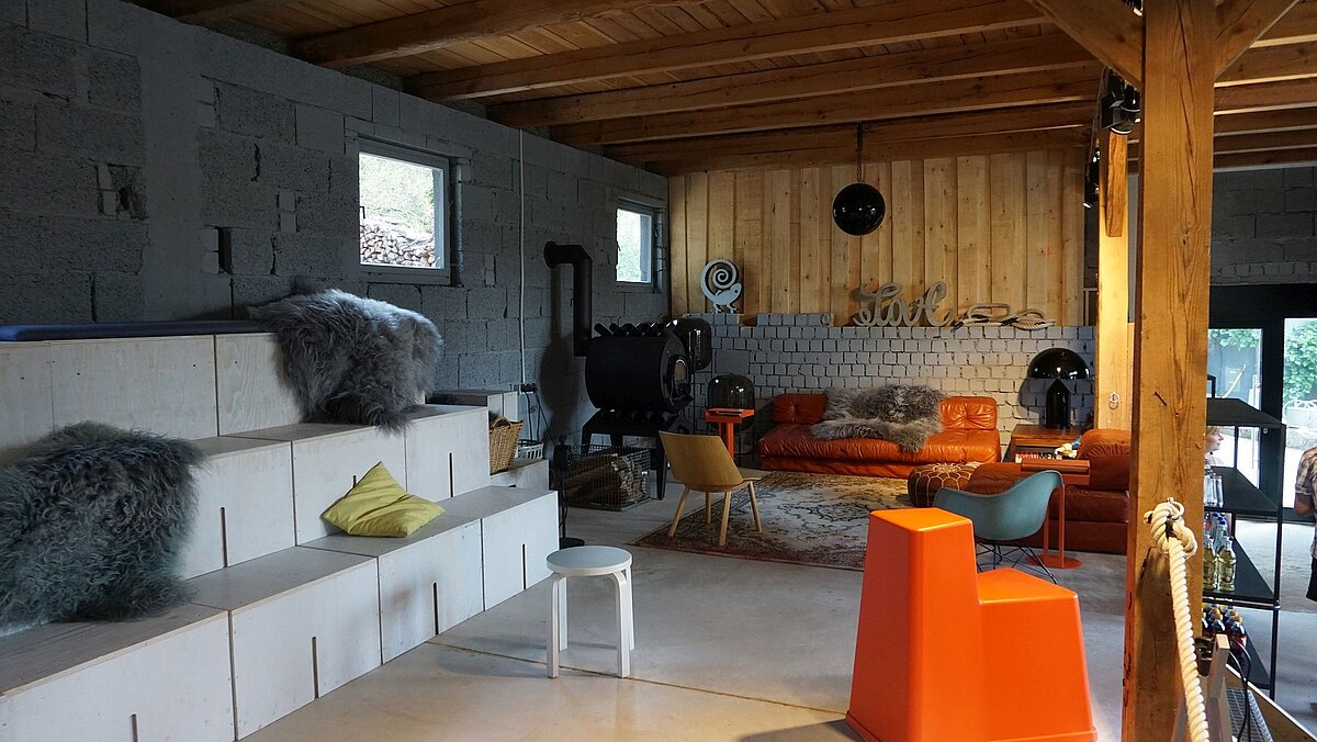 Wohnraum mit industriellem Flair: weiße Sitzstufen mit Kissen, Holzofen, orange Ledersofas, gemischte Möbel und Dekoelemente unter Holzbalkendecke in kreativ gestalteter Umgebung.
