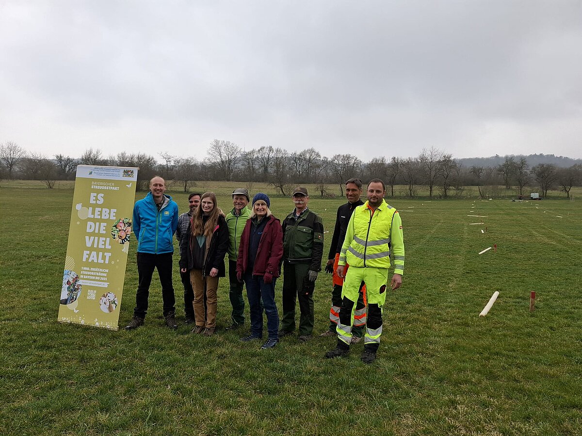 Acht Personen stehen auf einer Wiese neben einem gelben Banner mit der Aufschrift „Es lebe die Vielfalt“, im Hintergrund sind Bäume und ein bewölkter Himmel zu sehen, auf der Wiese liegen mehrere Holzpfosten und Markierungen.