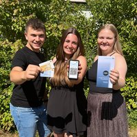 Drei junge Personen halten blaue und goldene Ehrenamtskarte, ein Smartphone und einen Flyer zur Ehrenamtskarte  in die Kamera