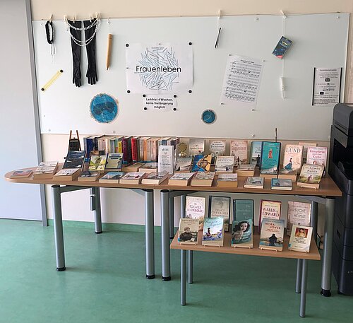 Zwei Tische mit zahlreichen Büchern in verschiedenen Formaten und Farben, dahinter ein Whiteboard mit dem Schriftzug „Frauenleben“ sowie aufgehängten Gegenständen wie Handschuhen, Perlenkette, Notenblättern und einem blauen Kreisdiagramm.