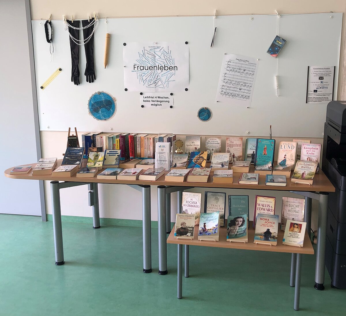 Zwei Tische mit zahlreichen Büchern in verschiedenen Formaten und Farben, dahinter ein Whiteboard mit dem Schriftzug „Frauenleben“ sowie aufgehängten Gegenständen wie Handschuhen, Perlenkette, Notenblättern und einem blauen Kreisdiagramm.