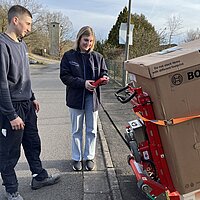 Zwei Personen stehen auf einer gepflasterten Straße neben einem großen Bosch-Karton auf einem roten Hebegerät – eine Person bedient das Gerät per Fernsteuerung, die andere beobachtet; im Hintergrund Bäume, Gebäude und Straßenlaterne.