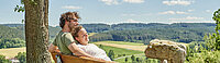 Ein stiller Moment auf der Bank am Hügel: Ein Pärchen genießen eng aneinandergeschmiegt den weiten Blick über Felder und Wälder – ein Bild voller Zärtlichkeit, Naturverbundenheit und friedlicher Gelassenheit.