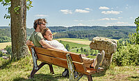 Ein stiller Moment auf der Bank am Hügel: Ein Pärchen genießen eng aneinandergeschmiegt den weiten Blick über Felder und Wälder – ein Bild voller Zärtlichkeit, Naturverbundenheit und friedlicher Gelassenheit.
