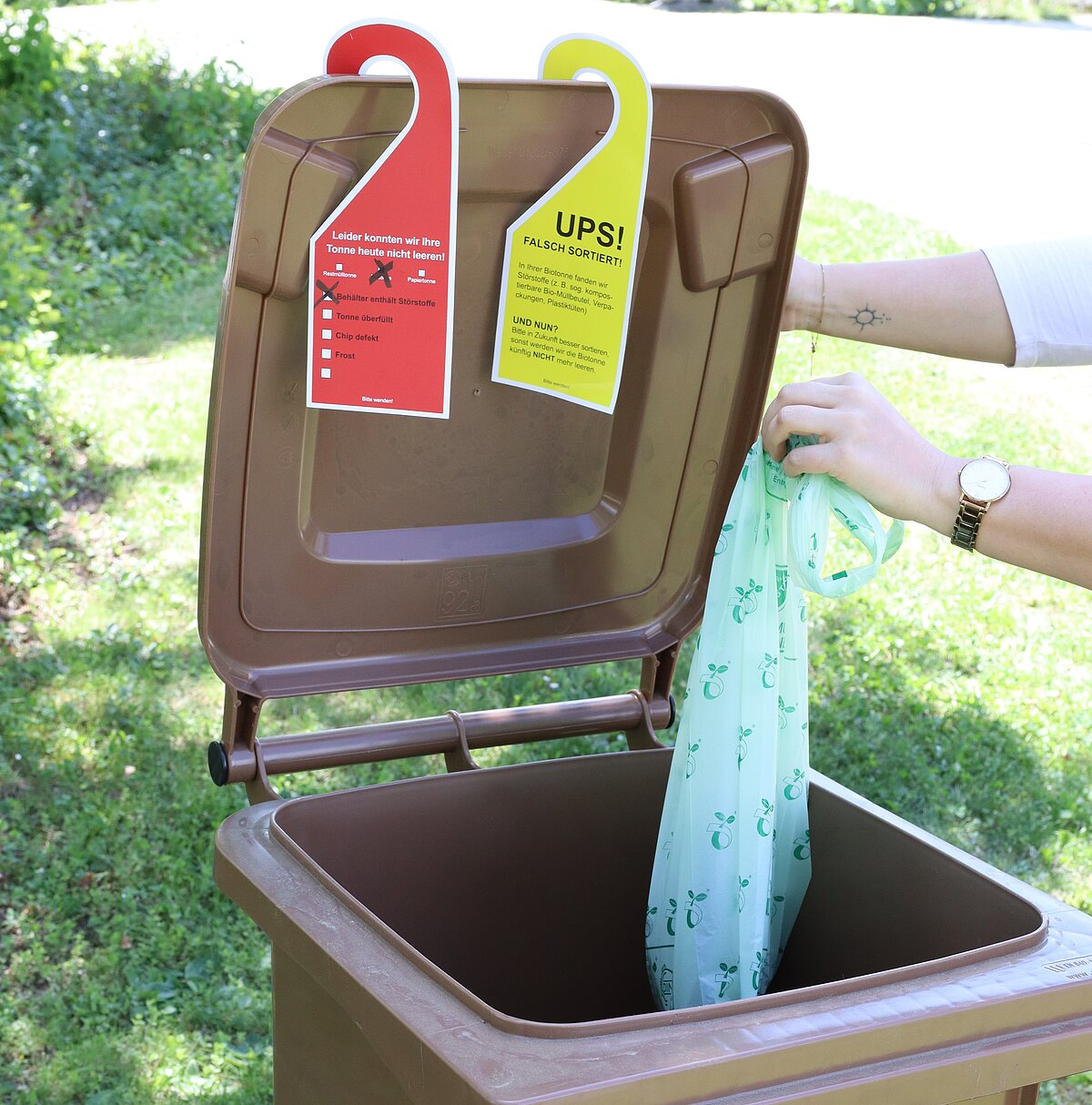 Eine Person wirft einen hellblauen Beutel mit grünem Muster in eine braune Mülltonne, deren geöffneter Deckel mit einem roten und einem grünen Hinweisanhänger zur Mülltrennung versehen ist.