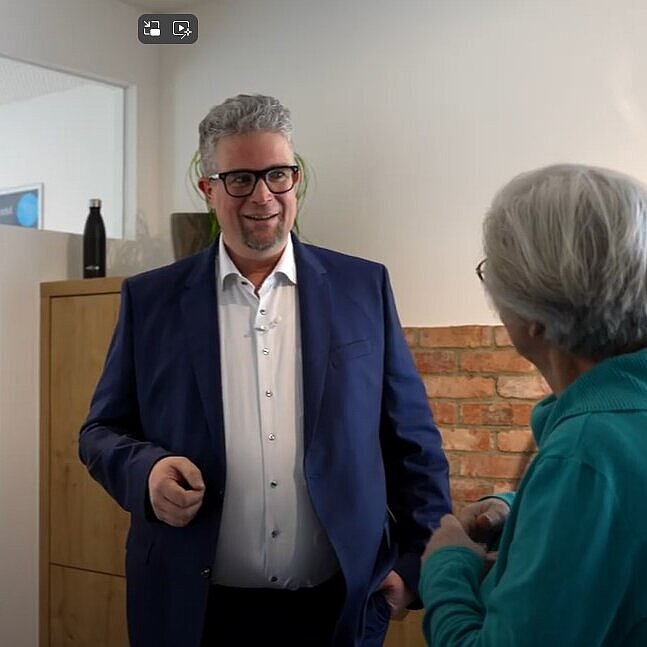 Drei Personen stehen in einem Innenraum vor einer Backsteinwand und unterhalten sich; eine Person im blauen Anzug spricht, die anderen hören zu – auf dem Tisch eine Flasche mit grünem Etikett, im Hintergrund ein Schrank und ein Poster.