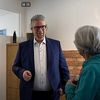 Drei Personen stehen in einem Innenraum vor einer Backsteinwand und unterhalten sich; eine Person im blauen Anzug spricht, die anderen hören zu – auf dem Tisch eine Flasche mit grünem Etikett, im Hintergrund ein Schrank und ein Poster.