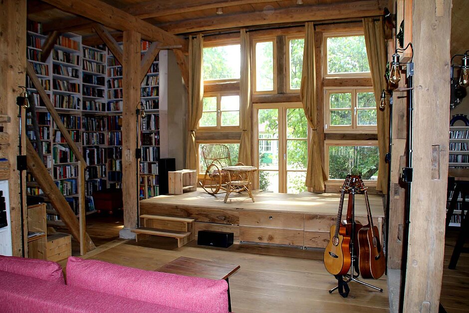 Fensterfront mit rechts und links Bücherregalen und vor dem Fenster eine kleine Bühne vor der ein rotes Sofa steht. 