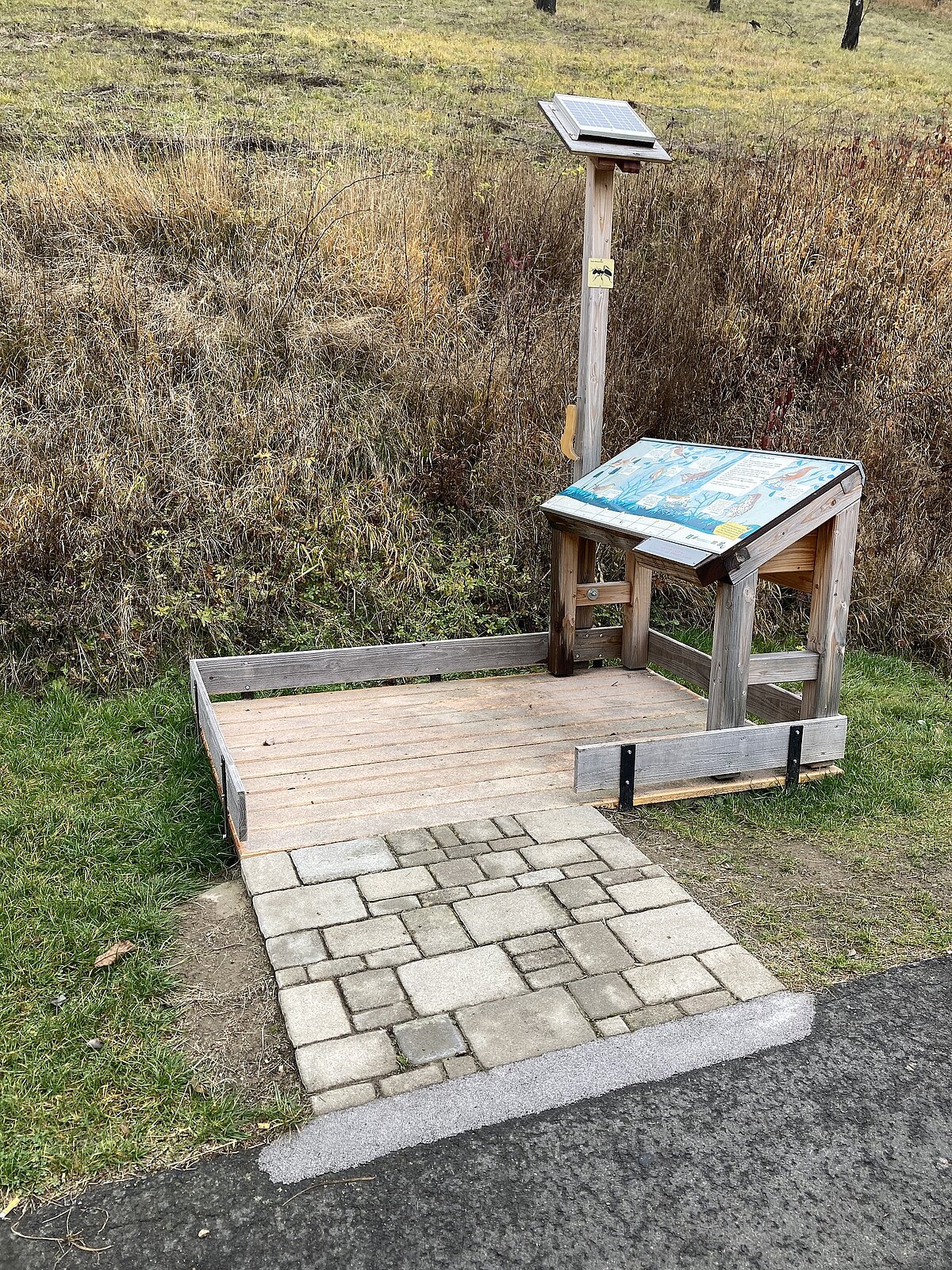 Eröffnung des barrierarmen Erlebnispfades in Marktbergel © Landratsamt Neustadt a.d.Aisch-Bad Windsheim Kleine Holzplattform mit solarbetriebenem Infopunkt am Wegesrand – ausgestattet mit geneigter Tafel unter Klarsichtabdeckung und Solarpanel auf Pfosten, umgeben von Pflasterweg, Holzrahmen und trockener Hangvegetation, Szene dient als naturnahe Informationsstelle in landschaftlicher Umgebung.