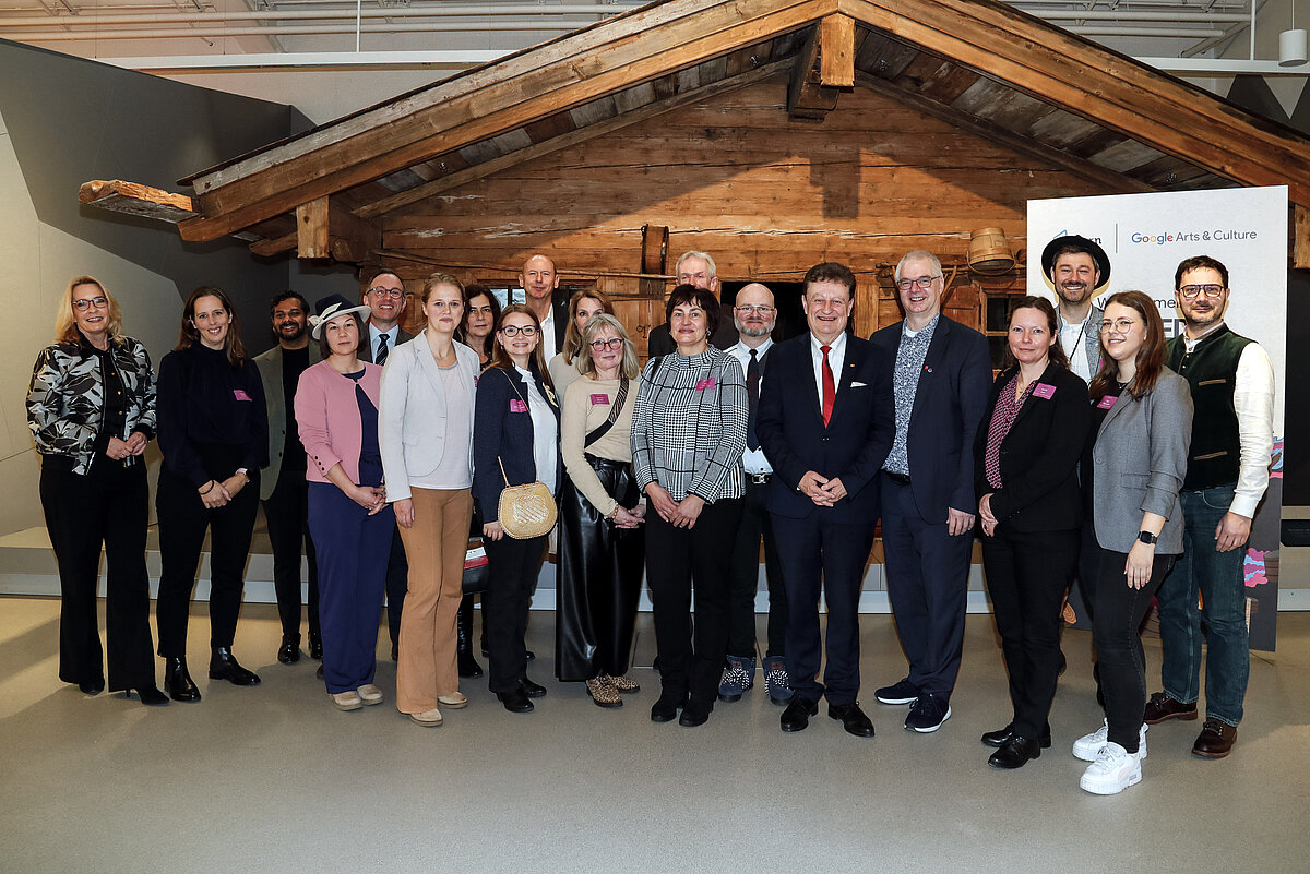 Gruppe von 20 Personen in Businesskleidung steht vor rustikaler Holzkulisse in Innenraum mit Banner „Google Arts & Culture“ – deutet auf kulturelle oder akademische Veranstaltung hin.