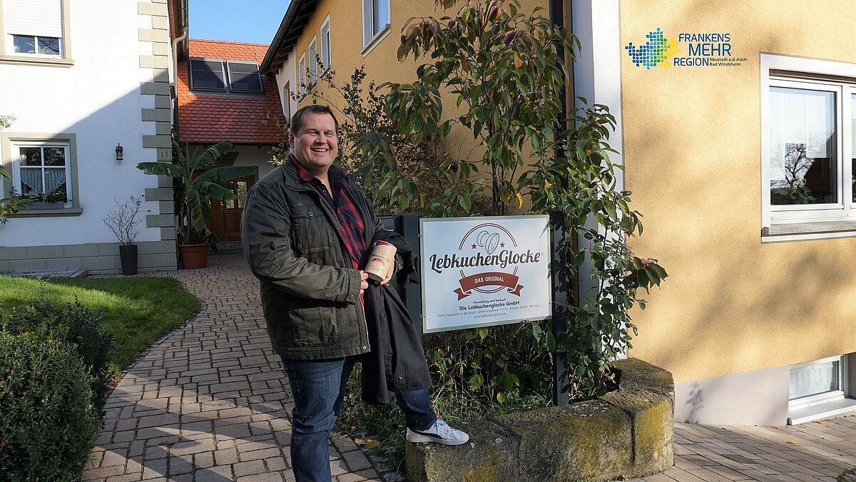 Ein herzliches Bild vor der „LebkuchenGlocke“: Ein lächelnder Besucher mit Gebäckdose in der Hand steht vor dem gelben Gebäude der Lebkuchenmanufaktur – ein süßer Gruß aus Franken, wo Tradition und Genuss Hand in Hand gehen.
