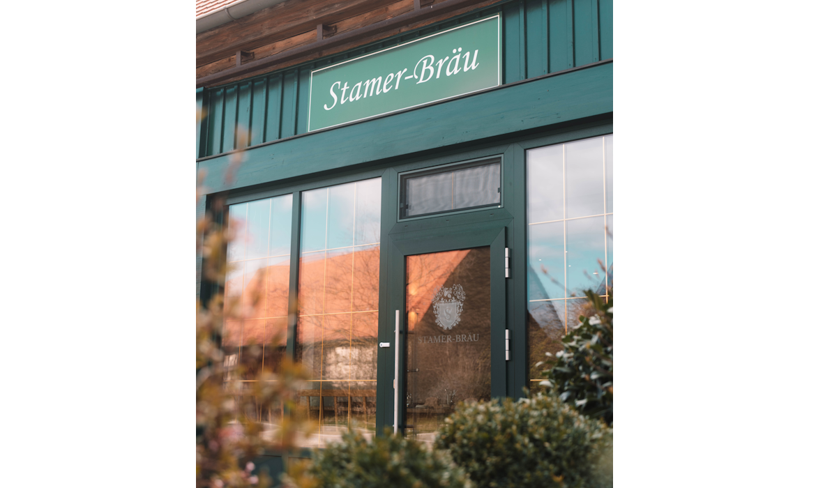 Außenansicht eines Gebäudes mit grüner Fassade und Glasfront – über dem Eingang das Schild „Stamer-Bräu“, ergänzt durch ein Logo mit Hopfenzapfen und Gerstenähren; die reflektierende Tür trägt ebenfalls den Schriftzug, im Vordergrund unscharfe Pflanzen.