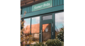 Außenansicht eines Gebäudes mit grüner Fassade und Glasfront – über dem Eingang das Schild „Stamer-Bräu“, ergänzt durch ein Logo mit Hopfenzapfen und Gerstenähren; die reflektierende Tür trägt ebenfalls den Schriftzug, im Vordergrund unscharfe Pflanzen.