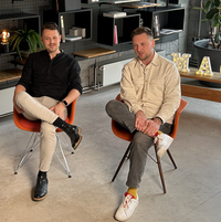 Zwei Personen sitzen auf orangefarbenen Stühlen in einem modern eingerichteten Raum mit Betonboden und großen Fenstern; hinter ihnen ein Regal mit Büchern und Dekoration, daneben eine Pflanze und ein Tisch.