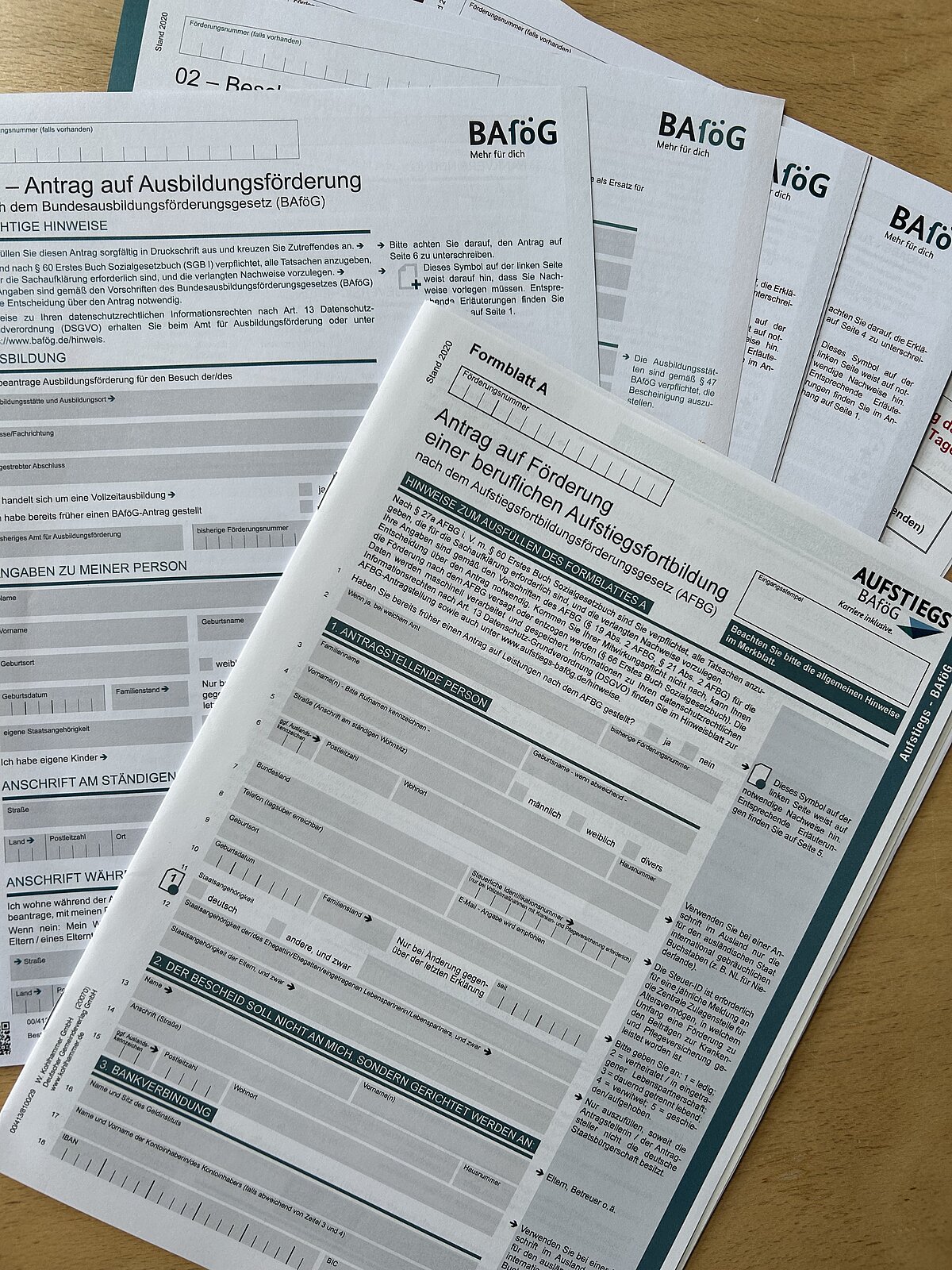 Mehrere BAföG-Formulare und Anträge auf Ausbildungsförderung sowie ein Antrag auf Förderung einer beruflichen Aufstiegsfortbildung liegen gestapelt auf einem Holztisch.