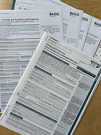 Mehrere BAföG-Formulare und Anträge auf Ausbildungsförderung sowie ein Antrag auf Förderung einer beruflichen Aufstiegsfortbildung liegen gestapelt auf einem Holztisch.
