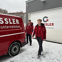 Zwei Personen in rot-schwarzer Arbeitskleidung gehen nebeneinander auf schneebedecktem Boden vor einem roten Transporter und einer Garage mit „Gessler Unternehmen“-Logo.