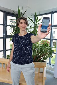 Eine Person steht in einem hellen Raum mit großen Fenstern und grünen Pflanzen im Hintergrund und hält ein Smartphone in der ausgestreckten Hand, auf dessen Display eine App mit blauen und weißen Bedienelementen sichtbar ist. Die Person trägt ein dunkelblaues T-Shirt mit weißen Punkten und eine helle Hose.