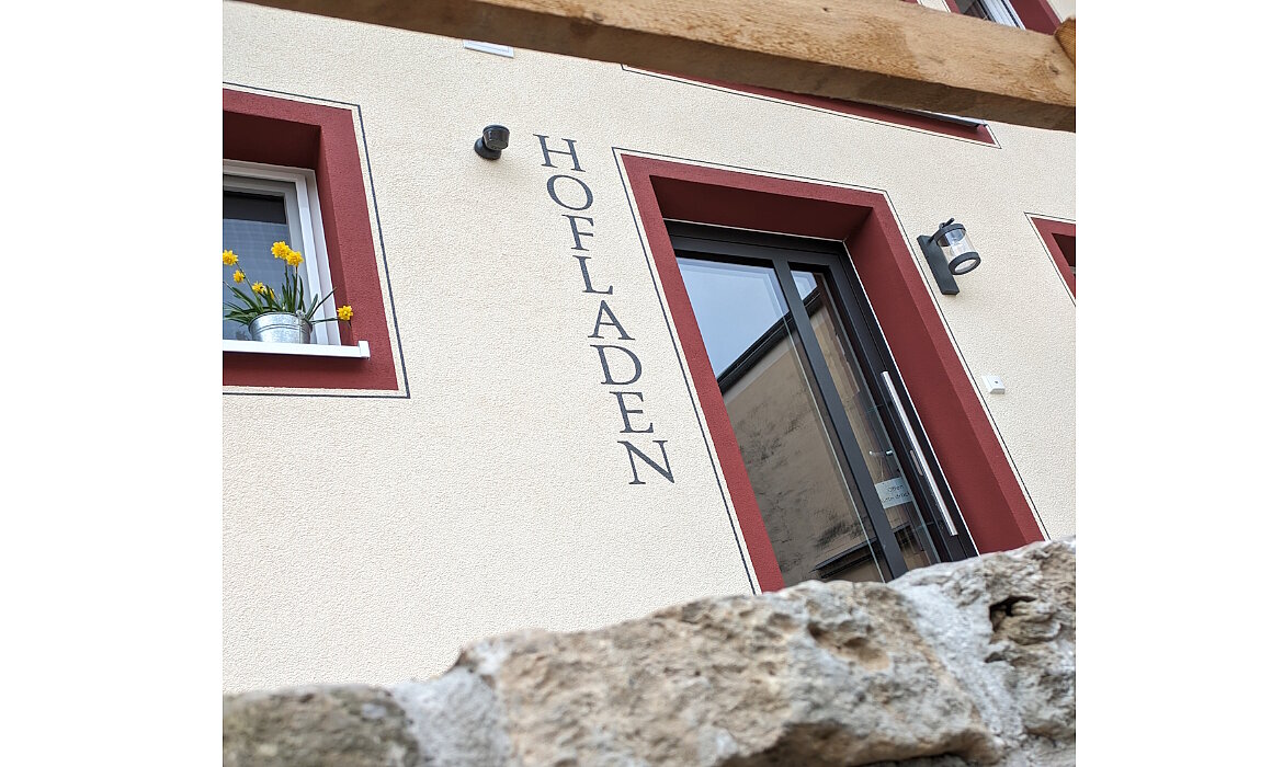 Einladender Eingang zum Hofladen: Mit roten Fensterrahmen, frischen Frühlingsblumen und dem Schriftzug „HOFLADEN“ an der Fassade strahlt das Gebäude ländliche Herzlichkeit und regionale Verbundenheit aus – ein Ort, an dem Qualität und Nähe zur Natur zuhause sind.