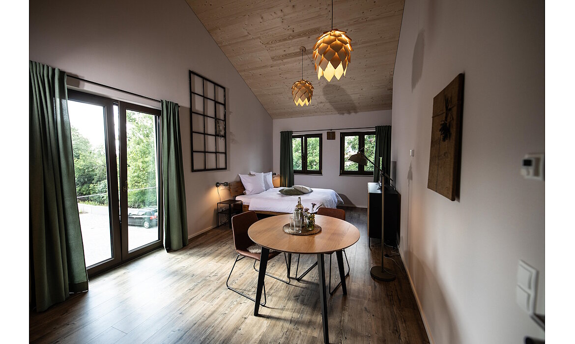 Modernes Schlafzimmer mit weiß bezogenem Bett vor drei Fenstern – Holzdecke und -boden, runde Sitzecke mit Tablett, grüne Vorhänge und kunstvolle Pendelleuchten schaffen ein ruhiges, stilvolles Ambiente mit Blick ins Grüne.