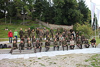 Gruppenfoto im Freien mit rund 50 Personen – Großteil trägt braune Uniformen, einige in farbiger Freizeitkleidung, Szene auf gestuften Sitzflächen vor Bäumen und Steinmauer, deutet auf gemeinschaftliche Veranstaltung oder Teamtreffen in naturnaher Umgebung.