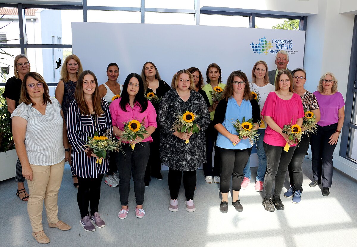 Gruppe von Personen steht in einem hellen Raum vor einer weißen Wand mit dem Logo „Frankens MEHR Region“, mehrere Personen halten Sträuße mit Sonnenblumen in den Händen; im Hintergrund sind große Fenster und Pflanzen sichtbar.