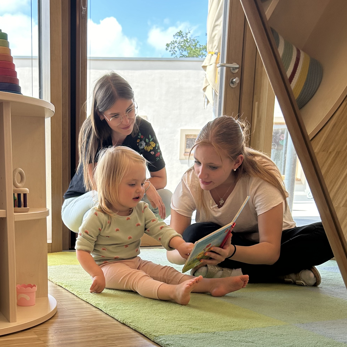 Drei Personen sitzen auf einem grünen Teppich in einem hellen Raum mit Holzregalen und großen Fenstern; ein Erwachsener liest einem Kind ein Buch vor, während die andere erwachsene Person lächelnd zusieht.