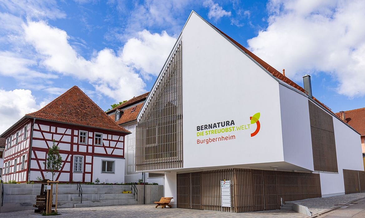 Modernes Gebäude mit markanter Dreiecksform und vertikaler Holzlattung trägt die Aufschrift „BERNATURA DIE STREUOBSTWELT Burgbernheim“ – daneben ein traditionelles Fachwerkhaus; gepflasterter Vorplatz mit Bank und gepflegter Umgebung betont den Kontrast zwischen zeitgenössischer Architektur und regionaler Baukultur.