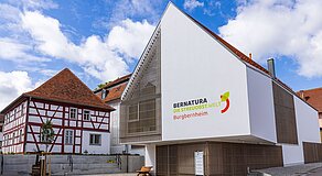 Modernes Gebäude mit markanter Dreiecksform und vertikaler Holzlattung trägt die Aufschrift „BERNATURA DIE STREUOBSTWELT Burgbernheim“ – daneben ein traditionelles Fachwerkhaus; gepflasterter Vorplatz mit Bank und gepflegter Umgebung betont den Kontrast zwischen zeitgenössischer Architektur und regionaler Baukultur.