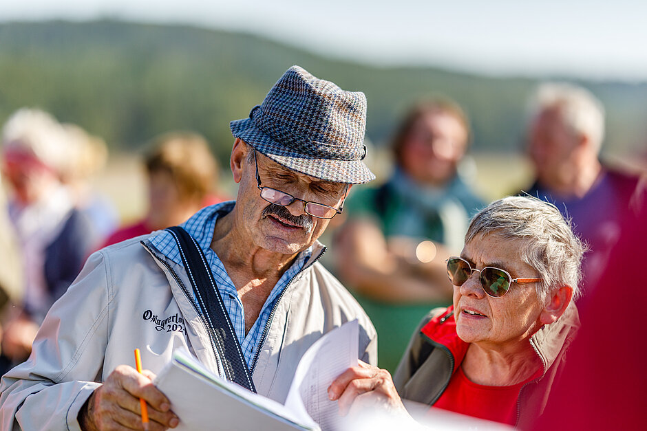 Eine Mann und eine Frau im Vordergrund die in Unterlagen sehen in der Natur mit Personen im Hintergrund