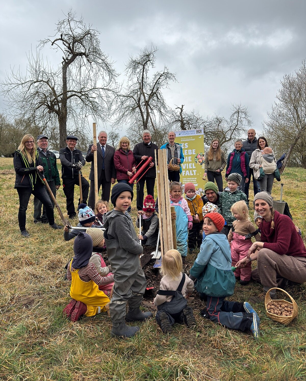 Gruppe von Erwachsenen und Kindern steht auf einer Wiese um eine frisch gepflanzte junge Pflanze mit Holzschutz, mehrere Kinder halten Werkzeuge, im Vordergrund ein Korb mit Pflanzmaterial, im Hintergrund ein gelbes Banner mit der Aufschrift „Es lebe die Vielfalt“ und kahle Bäume unter grauem Himmel.