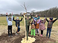 Kinder und eine Erwachsene Person stehen hinter einem frisch gepflanzten Obstbaum