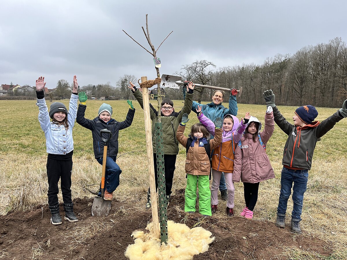 Kinder und eine Erwachsene Person stehen hinter einem frisch gepflanzten Obstbaum